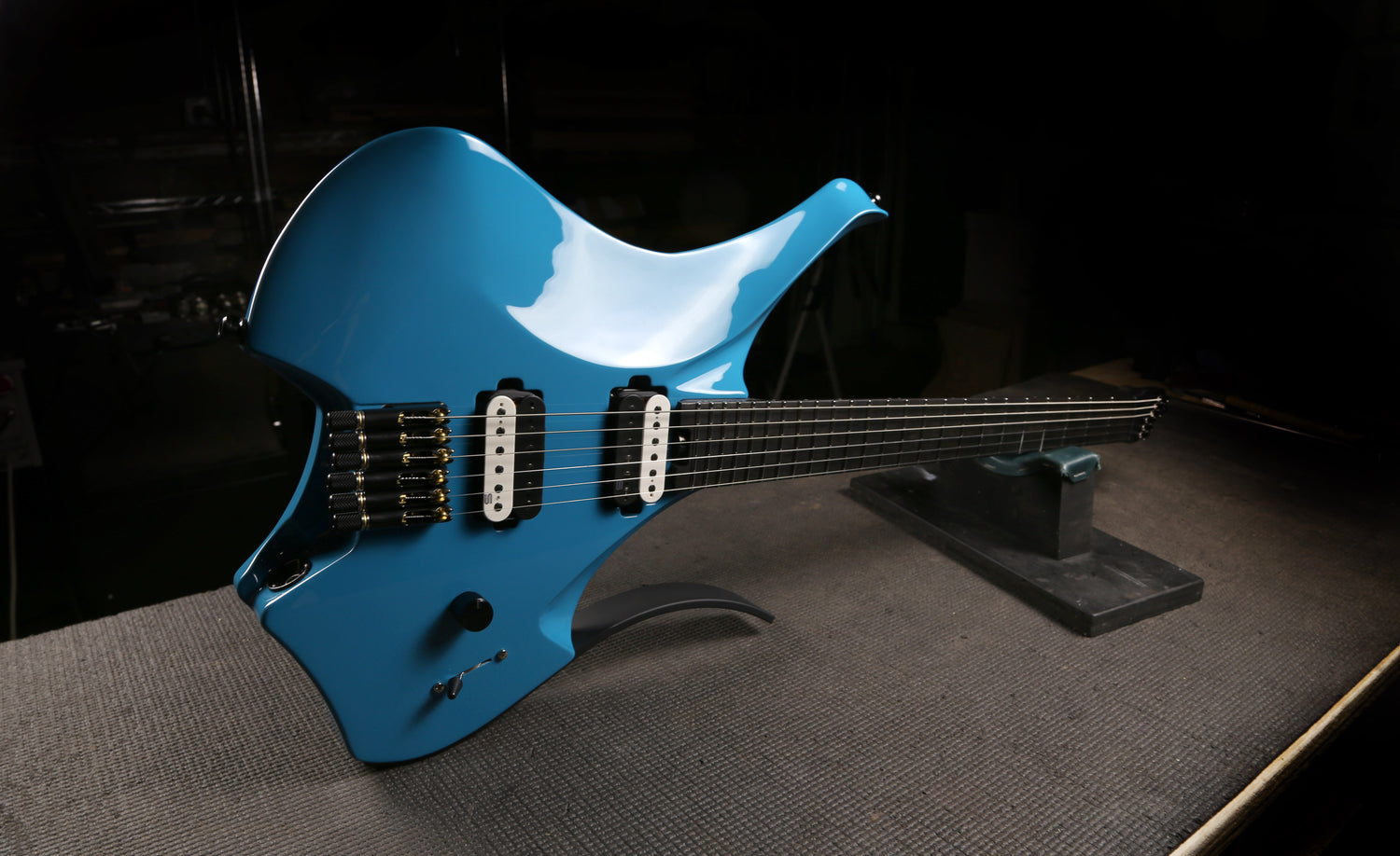 A bright blue electric guitar with a unique double-cutaway body, resting on a black stand on a textured gray surface against a dark background.